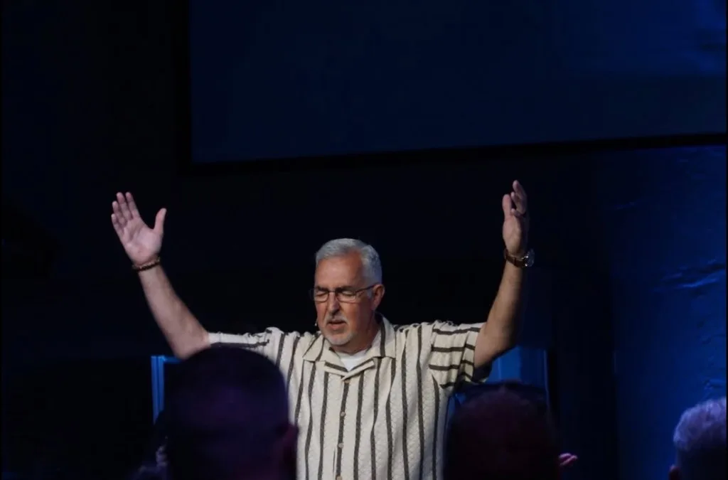 A worship leader with arms open wide before the congregation at The Refuge.
