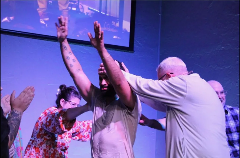 A man standing in the baptismal waters with hands raised as leaders pray over him at The Refuge.