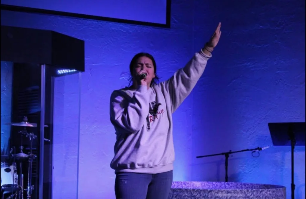 A young woman leading worship with her hand raised under blue stage light at The Refuge.