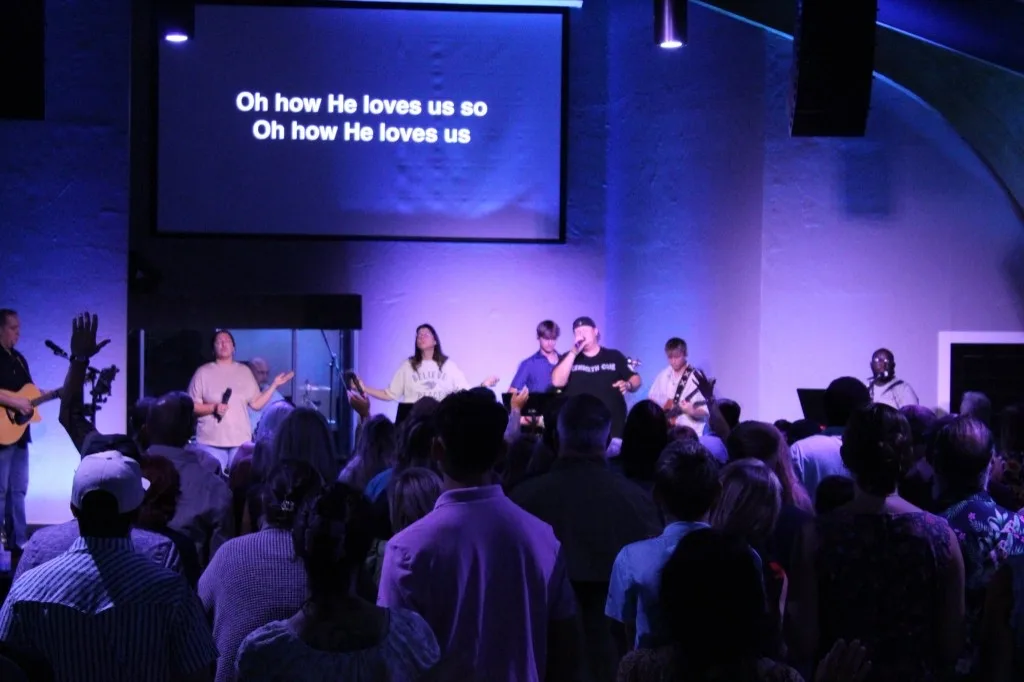 The Refuge worship team leading under purple light as the room sings “Oh how He loves us.”