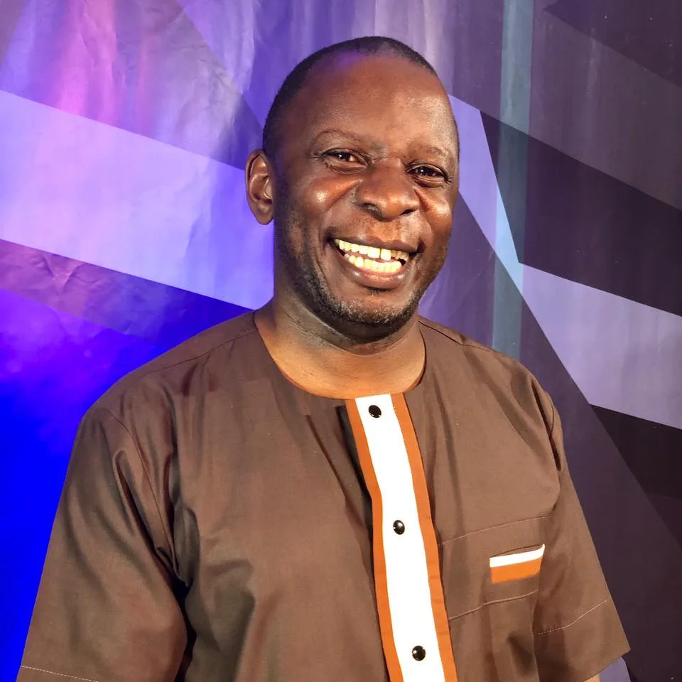 Pastor James Kato smiling in traditional attire against a purple backdrop.